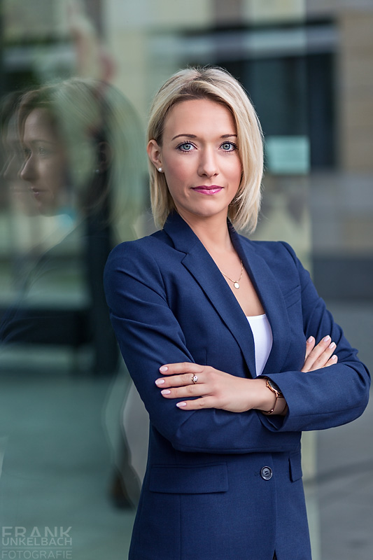 Business-Portrait einer attraktiven blonden Frau mit blauem Jacket und weißem Top. Schöne Spiegelung in einer Glasfasade.