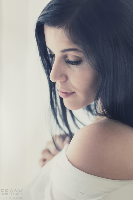 Romantisches Portrait einer schwarzhaarigen Frau in weißer Bluse vor einem Fenster. Sie schaut in Gedanken zur Seite und zeigt Ihr Schulter.