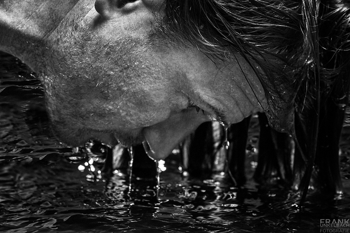 Ein attraktiver Mann mit Dreitagebart tauch aus dem Wasser auf. Wassertropfen fließen über Haare, Nase und Mund zurück ins kühle Nass.