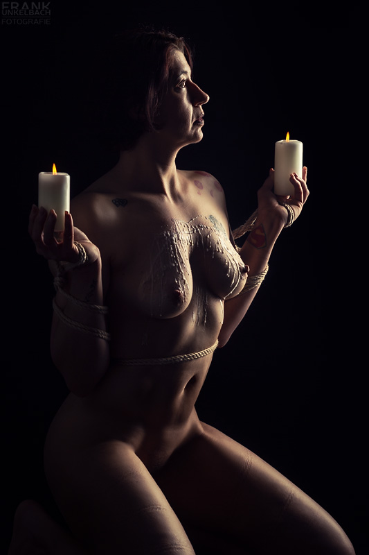 Mit einem Tengu gefesselte Frau kniet auf dem Boden und hält zwei Kerzen in der Hand. Auf Ihre Brüste wurde Wachs getropft.